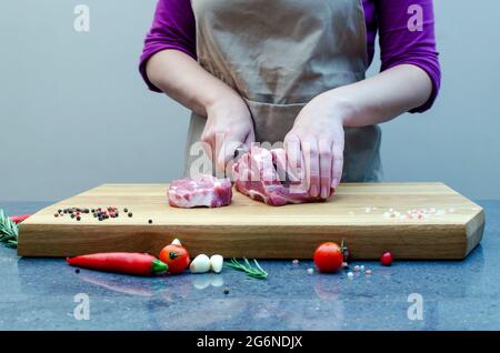 La padrona di casa in cucina trita il maiale con un coltello. Carne su un tagliere, preparazione di bistecca, preparazione per le vacanze. Spezie, peperoncino, rosmarino. Foto Stock