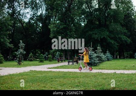 cartolina estiva, una ragazza guida uno scooter in un verde parco estivo con alberi e un prato verde sullo sfondo Foto Stock