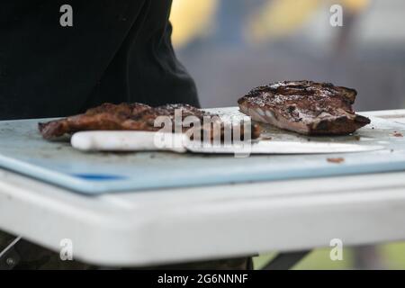 Un uomo che taglia le costole del barbecue dalla griglia fumatore sul tagliere con i guanti sopra e un coltello grande. Foto Stock