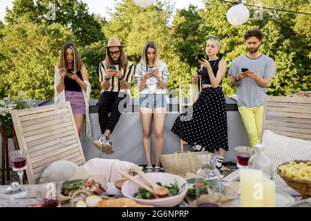 Le persone trascorrono il tempo insieme, seduti separatamente in smartphone durante le feste all'aperto Foto Stock