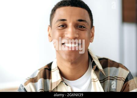 Positivo gara mista giovane uomo. Primo piano di un bel ragazzo latino millenario soddisfatto con, freelance, designer creativo o studente, in piedi in casa, guardando direttamente la fotocamera, sorridente amichevole Foto Stock