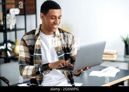 Positivo sicuro moderno giovane razza mista uomo latino con occhiali, designer creativo o freelance, elegantemente vestito, stare vicino a una scrivania con portatile aperto, guarda lo schermo, sorrisi, naviga in internet Foto Stock