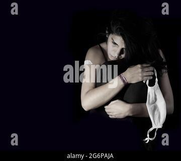 Bruna ragazza seduta abbracciando le gambe molto stressata dopo il vaccino del coronavirus, con un cerotto sul braccio, un bracciale e una maschera in mano. Horizo Foto Stock