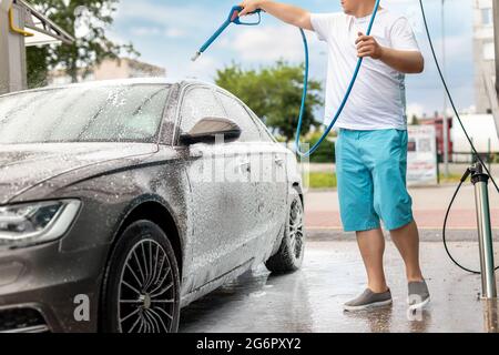 Primo piano dettaglio autolavaggio con pompa ad acqua ad alta pressione all'aperto self-service nella brillante giornata estiva. Veicolo coperto con shampoo in schiuma Foto Stock