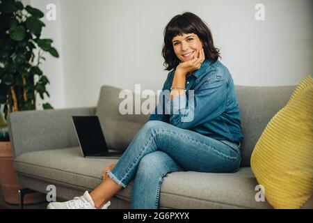 Donna sul divano nell'atrio che guarda la macchina fotografica e sorride. Donna sicura che sorride alla macchina fotografica mentre si siede sul divano in ufficio. Foto Stock
