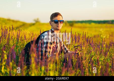 Giovane uomo in occhiali da sole seduto in un campo con fiori selvatici, uomo in campagna durante il tramonto, godendo della natura, concetto di viaggio, natura Foto Stock