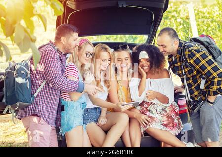 Gruppo di migliori amici multirazziali seduti sul bagagliaio dell'auto che si divertono veramente utilizzando il telefono cellulare in viaggio Foto Stock