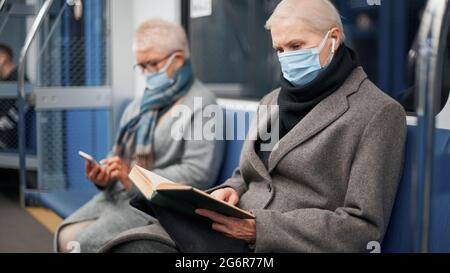 donna matura in maschera protettiva leggere un libro seduto in una metropolitana auto. regole della città durante la pandemia. Foto Stock