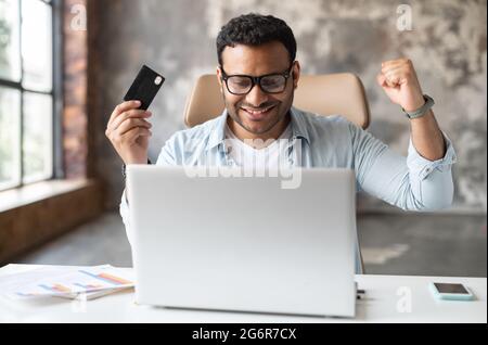 Un ragazzo misto-corsa fatto a lungo atteso acquisto online, seduto alla scrivania e sollevando pugni in gesto trionfale, tenendo la carta di credito, indiano uomo guarda lo schermo del portatile e gioisce Foto Stock