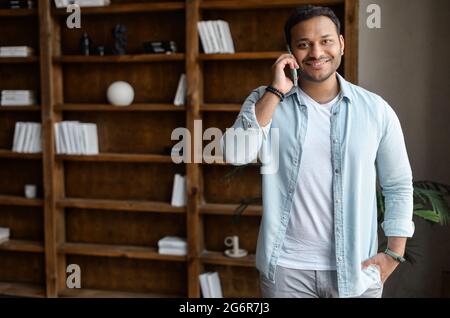 Un ragazzo indiano allegro e positivo in camicia casual che parla dallo smartphone nell'ufficio moderno. Felice uomo di razza mista ha conversazione telefonica. Gioioso dipendente maschile che parla al chiuso con il telefono cellulare Foto Stock