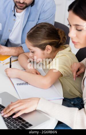 ragazza che scrive in notebook vicino a papà e madre offuscata utilizzando il computer portatile Foto Stock
