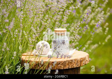 Vari prodotti di fiori di lavanda fatti in casa bomba da bagno e sale da bagno su vassoio di legno di pino naturale in campo di lavanda all'aperto in estate. Foto Stock
