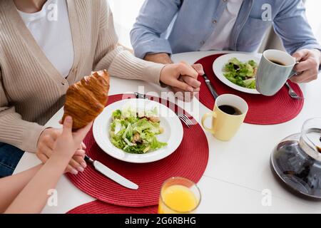 vista tagliata della ragazza che tiene croissant vicino ai genitori e insalata di verdure fresche Foto Stock