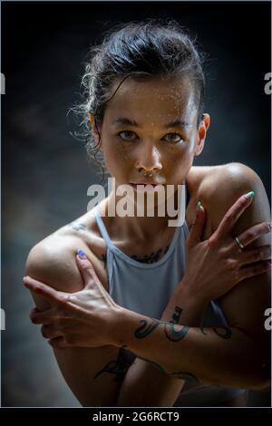 Una giovane donna con tatuaggi in abbigliamento sportivo, si sta abbracciando con le braccia sul petto. Sta guardando la macchina fotografica. Foto Stock