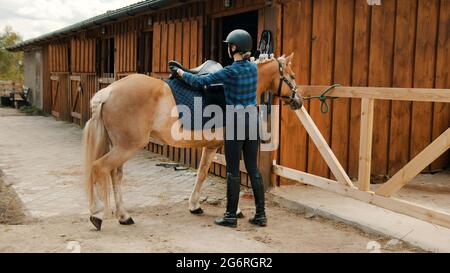 Jockey femminile che saddling il suo cavallo marrone chiaro fuori della scuderia. Disponendo una sella di cuoio sulla parte posteriore del suo cavallo. La ragazza che indossa un casco e guanti. Preparare il suo cavallo per la concorrenza. Foto Stock