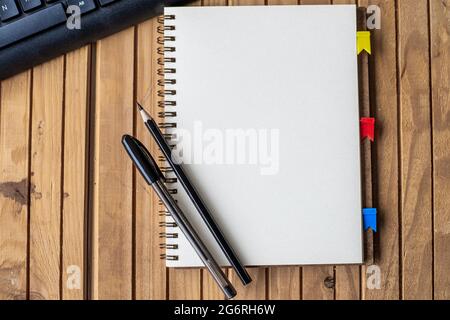 Giornale tagliato vuoto aperto accanto A una tastiera Pens sulla parte superiore della scrivania in legno. Blocco note vuoto aperto con clip e matite vicino alla tastiera posizionati sul tavolo. Foto Stock