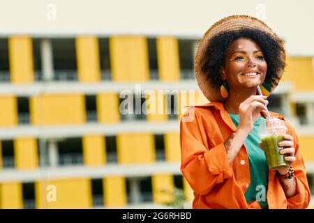 Bella donna afroamericana positiva con un frullato sano nelle sue mani sta camminando in una città urbana moderna Foto Stock