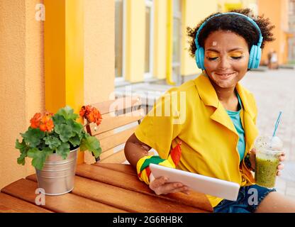 Donna giovane positiva con vitiligo che indossa cuffie blu e abiti alla moda si siede in un caffè estivo all'aperto e beve un cocktail vitaminico che guarda Foto Stock