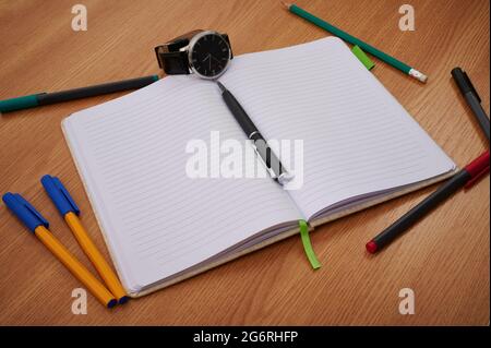 Orologio da polso per notebook vuoto circondato da assortimento di articoli per cancelleria. Svuotare Apri blocco note e verifica con Messy Office Pens Supply Over Foto Stock