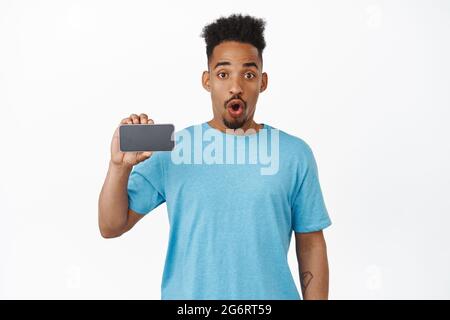 App fantastica dai un'occhiata. Stupito afro-americano guy gasp, dire WOW, mostrando orizzontale smartphone schermo, telefono cellulare interfaccia applicazione, staring Foto Stock