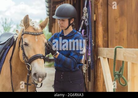 Jockey femminile con il suo cavallo marrone chiaro fuori dalla stalla. La ragazza è vestita con una camicia blu controllata e indossa un casco di sicurezza. Fissare la briglia del suo cavallo. Prepararsi alla concorrenza. Foto Stock