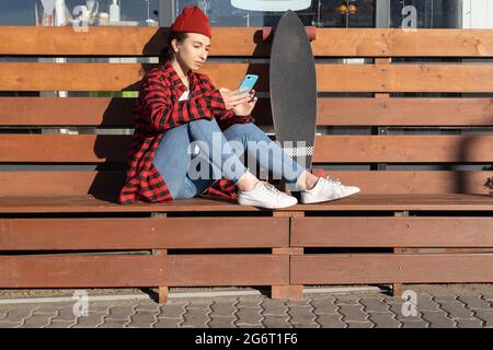 Ragazza hipster casual alla moda con smartphone seduto su panca all'aperto con longboard in piedi vicino Foto Stock