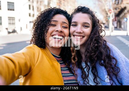 Belle ragazze giovani con capelli ricci sorridenti e prendendo selfie in strada. Foto Stock