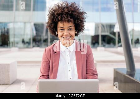 Donna di affari con i capelli afro che siede all'aperto nell'area di affari usando il suo calcolatore e guardando la macchina fotografica. Foto Stock