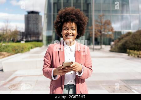 Donna di affari nera graziosa con i capelli afro usando il suo telefono mobile e guardando la macchina fotografica. Sta camminando in una zona di affari della città. Foto Stock