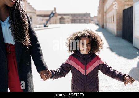 Adorabile giovane ragazza nera con capelli afro che cammina con la madre e tiene le mani. Sorride e guarda la macchina fotografica. Foto Stock