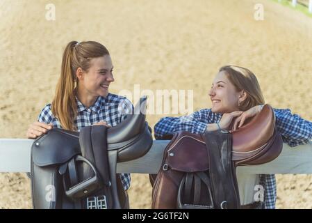 Due giovani ragazze in piedi con le mani poggiate sulla recinzione di legno nel ranch di cavalli. Ragazze di buon umore che ridono e parlano. Selle in pelle appese sulla recinzione in legno in primo piano. Foto Stock