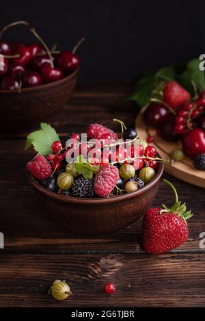 Fragole, ciliegie, ribes, lamponi, frutti di bosco in una ciotola di argilla su un tavolo di legno scuro, frutti di bosco sani. Foto Stock