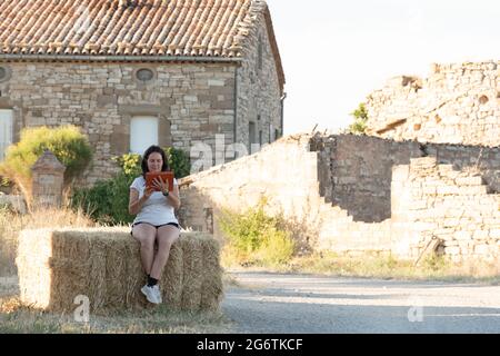 Scena rurale con bruna latina di mezza età donna consultando una tavoletta con coperchio arancione seduta su una balla di paglia con casa in pietra sullo sfondo. Foto Stock