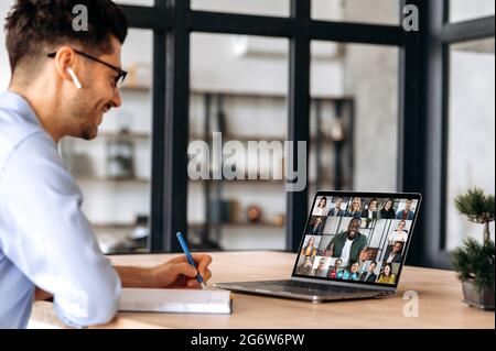 Un manager caucasico di successo, prende appunti durante la riunione di business online con colleghi multinazionali, utilizzando il laptop, seduto in ufficio, pianificazione strategia di marketing, colleghi di ascolto Foto Stock