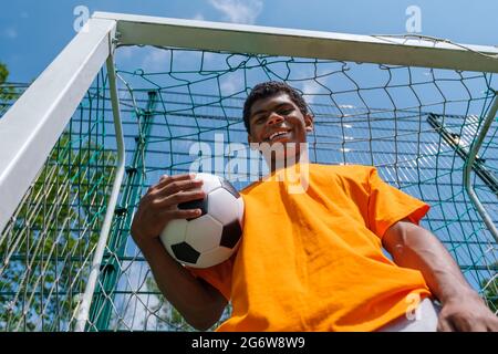 Uomo afroamericano sorridente che tiene la palla di calcio mentre si trova in piedi nel gol di calcio Foto Stock