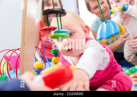 Carino bambina che mostra il progresso e curiosità cercando di raggiungere multicolore di giocattoli di legno, mentre è seduto sul pavimento con la madre e una famiglia ven Foto Stock