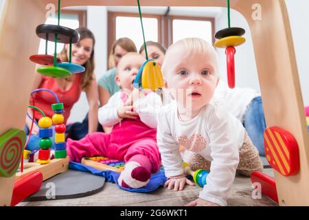 Carino bambina che mostra il progresso e curiosità cercando di raggiungere multicolore di giocattoli di legno, mentre è seduto sul pavimento con la madre e una famiglia ven Foto Stock