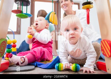 Carino bambina che mostra il progresso e curiosità cercando di raggiungere multicolore di giocattoli di legno, mentre è seduto sul pavimento con la madre e una famiglia ven Foto Stock