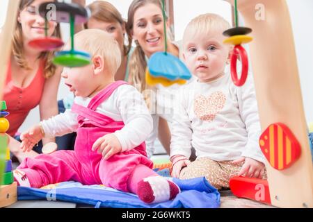 Carino bambina che mostra il progresso e curiosità cercando di raggiungere multicolore di giocattoli di legno, mentre è seduto sul pavimento con la madre e una famiglia ven Foto Stock