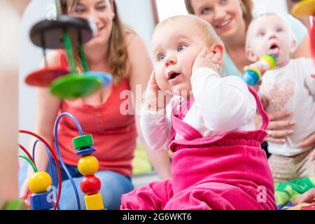 Carino bambina che mostra il progresso e curiosità cercando di raggiungere multicolore di giocattoli di legno, mentre è seduto sul pavimento con la madre e una famiglia ven Foto Stock