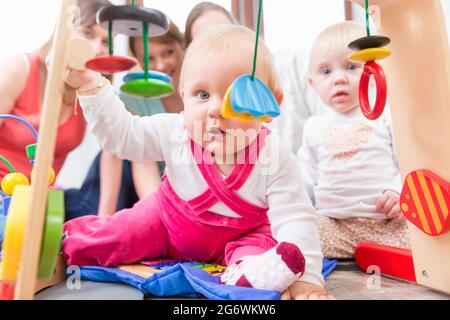 Carino bambina che mostra il progresso e curiosità cercando di raggiungere multicolore di giocattoli di legno, mentre è seduto sul pavimento con la madre e una famiglia ven Foto Stock