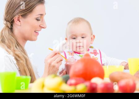 Ritratto di una bambina carina e sana, guardando con curiosità alla purea di frutta rosa mentre si siede su una sedia alta durante il pasto con la sua mamma felice Foto Stock