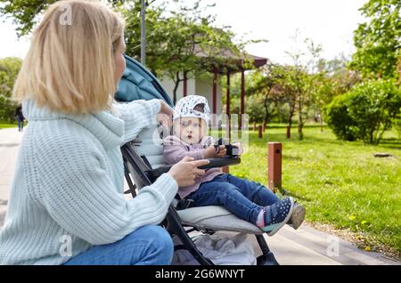 Mamma e figlia sulla natura a piedi presso il parco primaverile. Adorabile bambina in abiti comuni seduta in carrozzina blu. Bambino in buggy Foto Stock