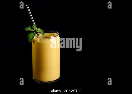 Mango lassi in vetro con tubo su fondo nero. Bevanda tradizionale indiana e pakistana con yogurt, mango, spezie, menta e ghiaccio Foto Stock