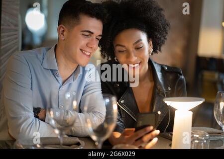 Una coppia di giovani interrazziali seduti su un tavolo da ristorante e guardando i contenuti dei social network sullo smartphone. Persone che utilizzano il concetto di tecnologia. Foto Stock