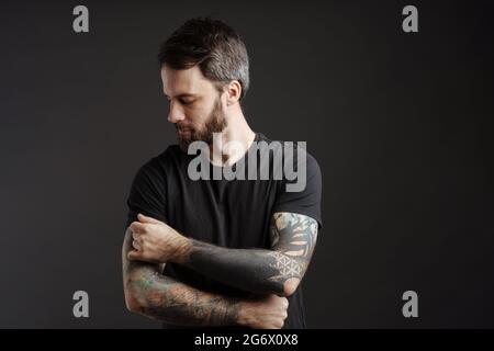Uomo bruna bearato con tatuaggio in posa e guardando verso il basso isolato su sfondo nero Foto Stock