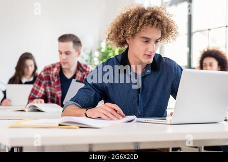 Ritratto di un giovane studente che lavora sodo seduto alla scrivania mentre scrive durante la lezione in un'università moderna Foto Stock