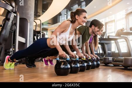 Vista laterale di tre giovani che sorridono mentre fanno la sfida kettlebell Plank per il nucleo forte durante l'allenamento di gruppo in palestra Foto Stock