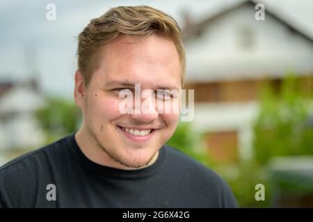 Un giovane sorridente e amichevole con una barba corta e spionata che grondola felicemente alla fotocamera in un primo piano ritratto all'aperto Foto Stock