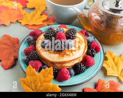 Colazione con snack accogliente con una pila di frittelle e frutti di bosco con tè verde con decorazioni in foglie autunnali Foto Stock